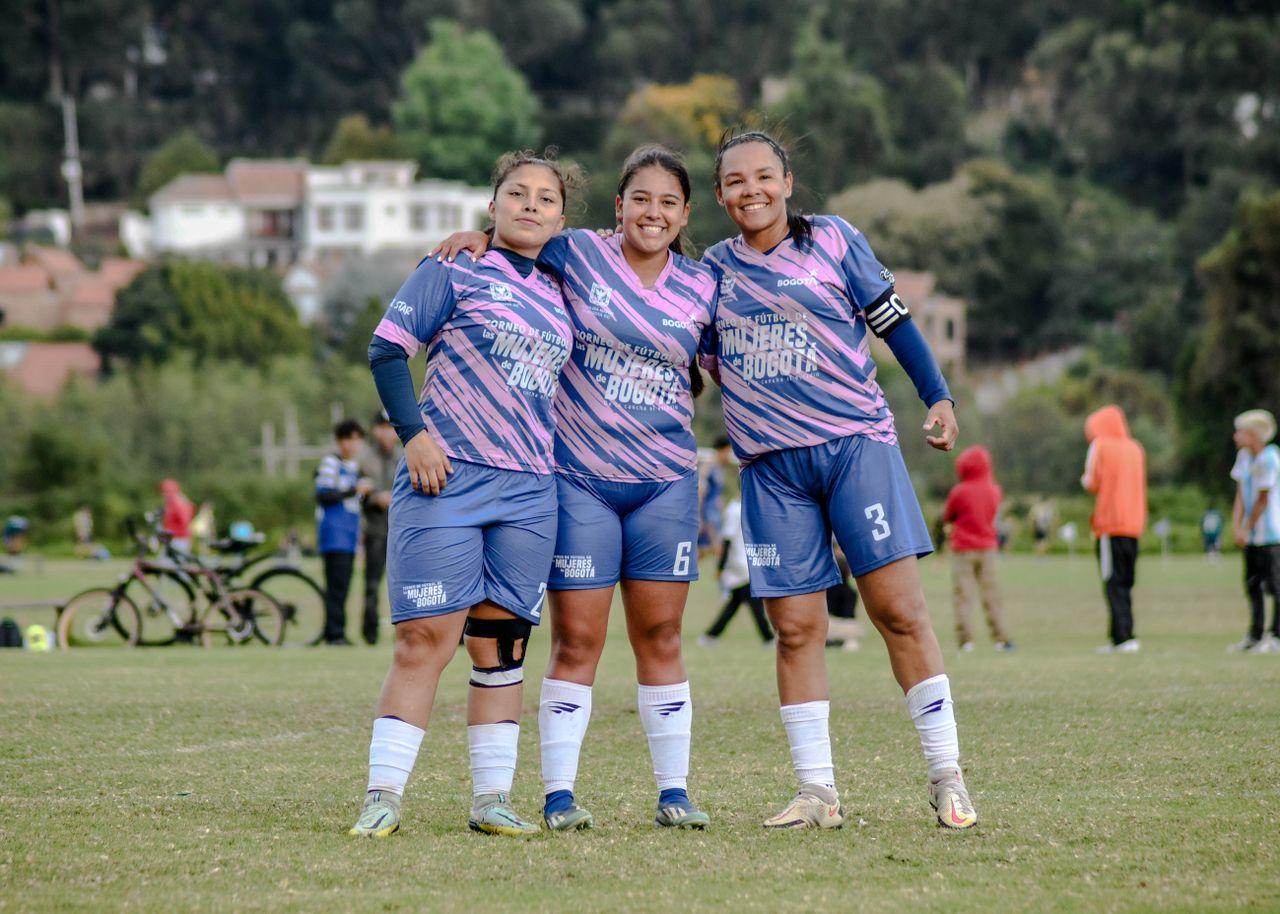 Three female soccer players in team uniform pose happily on a grass field.