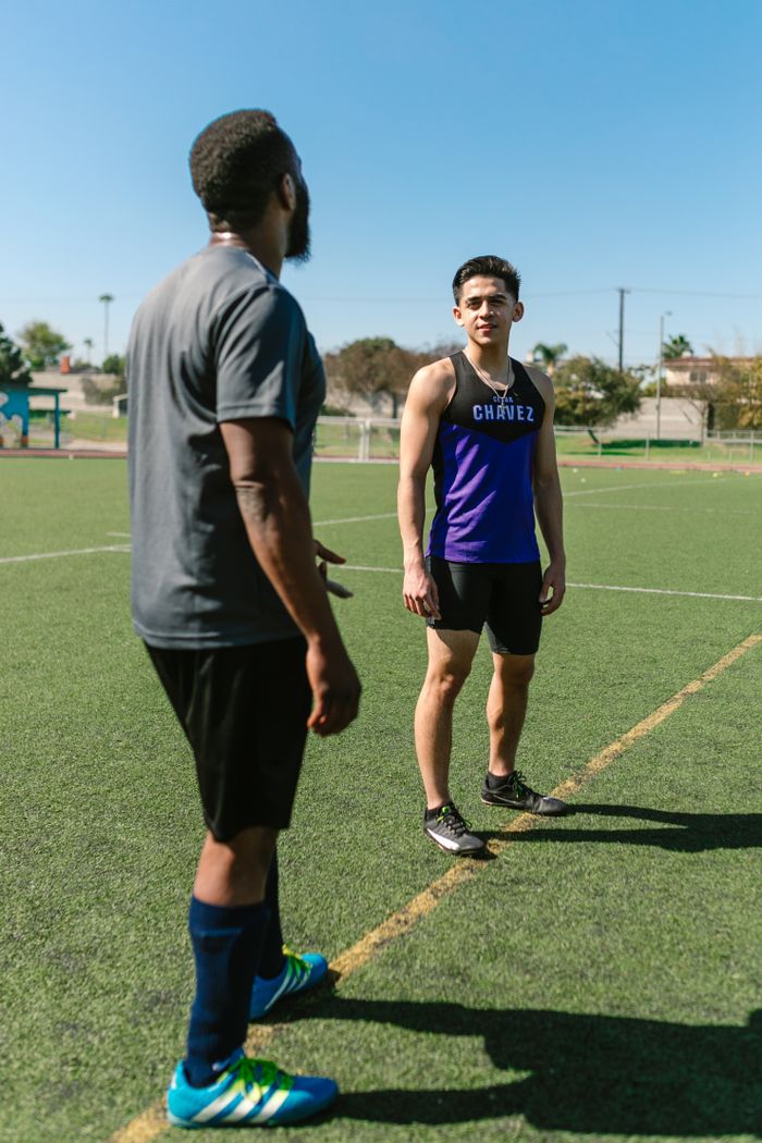 contact-img Two male athletes talking on an outdoor sports field during a sunny day, preparing for a workout.